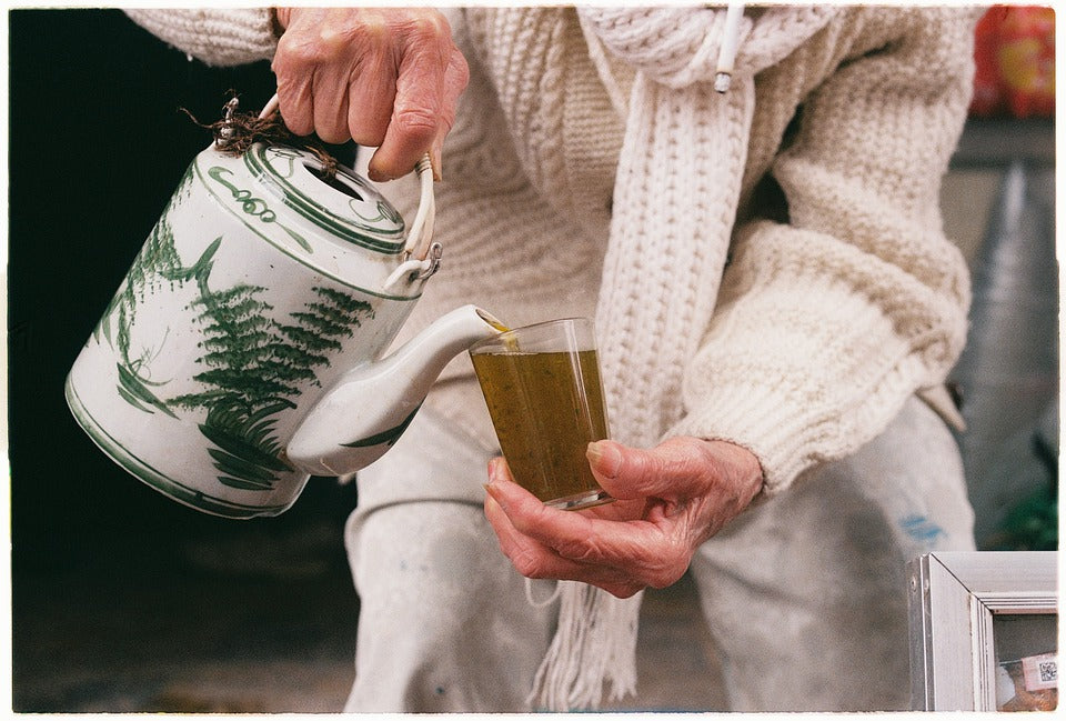 Older woman pouring loose green tea into a glass from an oriantal teapot decorated with green ferns