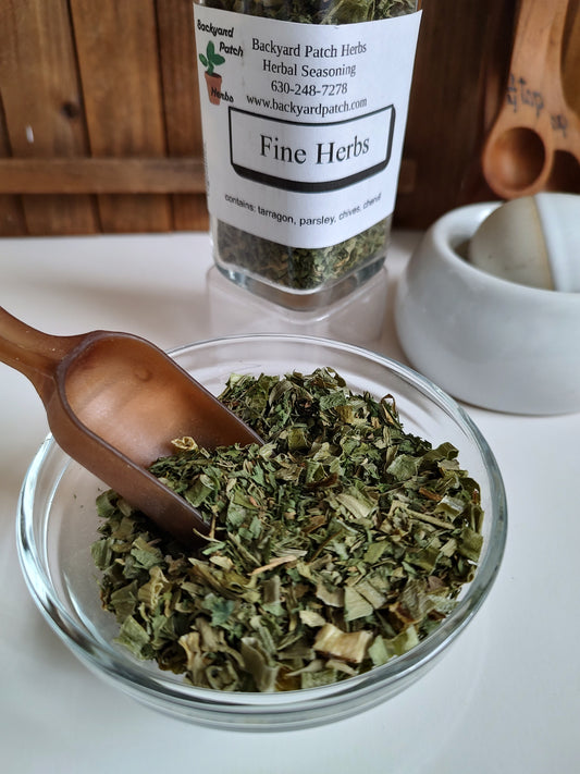 'Fine Herbs' seasoning in a glass jar with a wooden scoop on a dish of dried herbs on a white surface