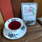 Tea cup with floral design filled with red brewed Summer Breese Tea next to a package of 'Summer Breeze Tea Blend' on a wooden surface.