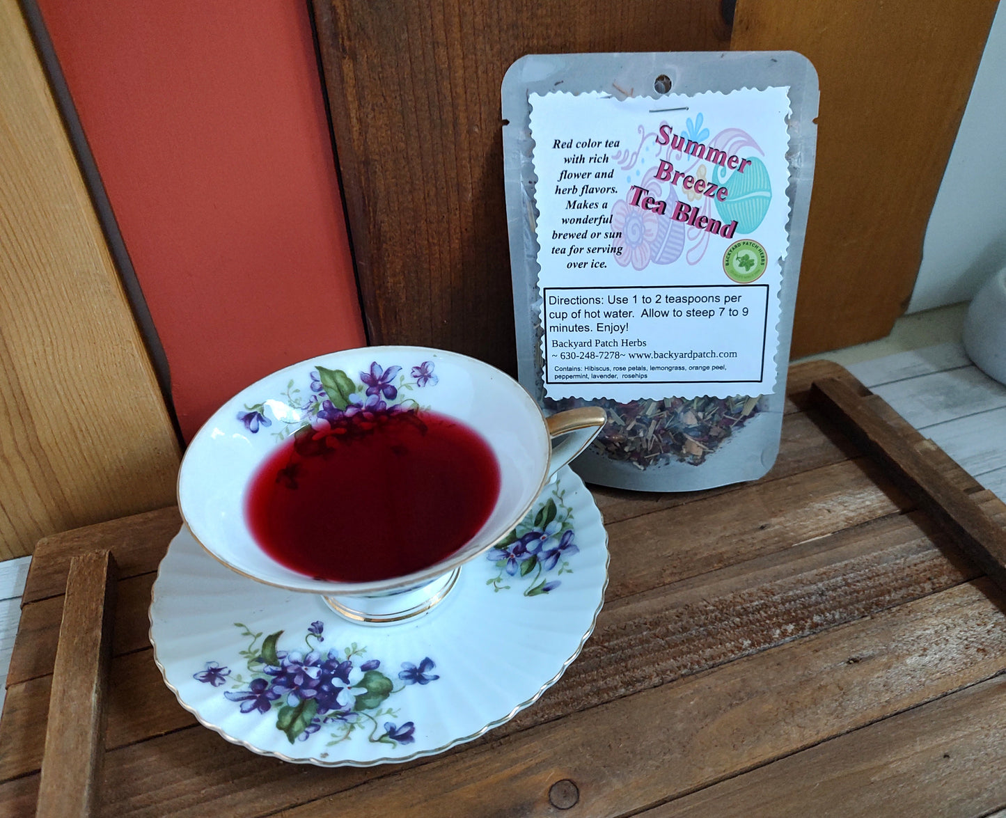 Tea cup with floral design filled with red brewed Summer Breese Tea next to a package of 'Summer Breeze Tea Blend' on a wooden surface.