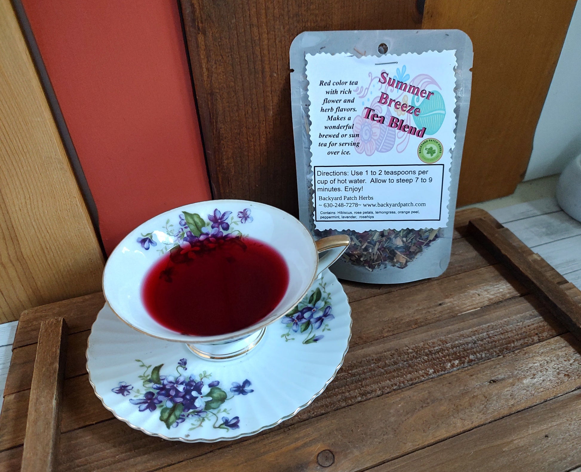 Tea cup with floral design filled with red brewed Summer Breese Tea next to a package of 'Summer Breeze Tea Blend' on a wooden surface.