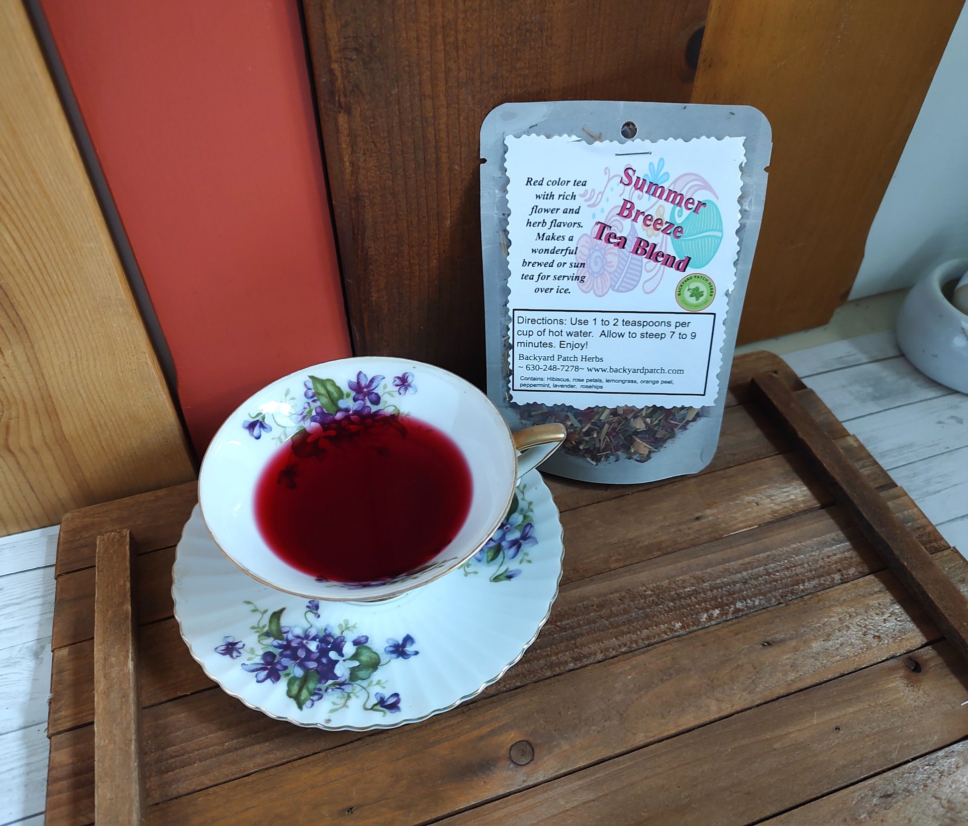 Tea cup with floral design filled with red brewed Summer Breese Tea next to a package of 'Summer Breeze Tea Blend' on a wooden surface.