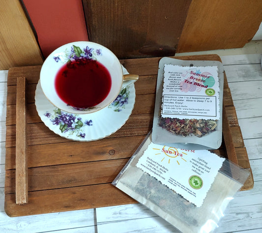 Tea cup with floral design filled with red brewed Summer Breeze Tea next to a package of loose  'Summer Breeze Tea Blend' and sun tea bags on a wooden surface.